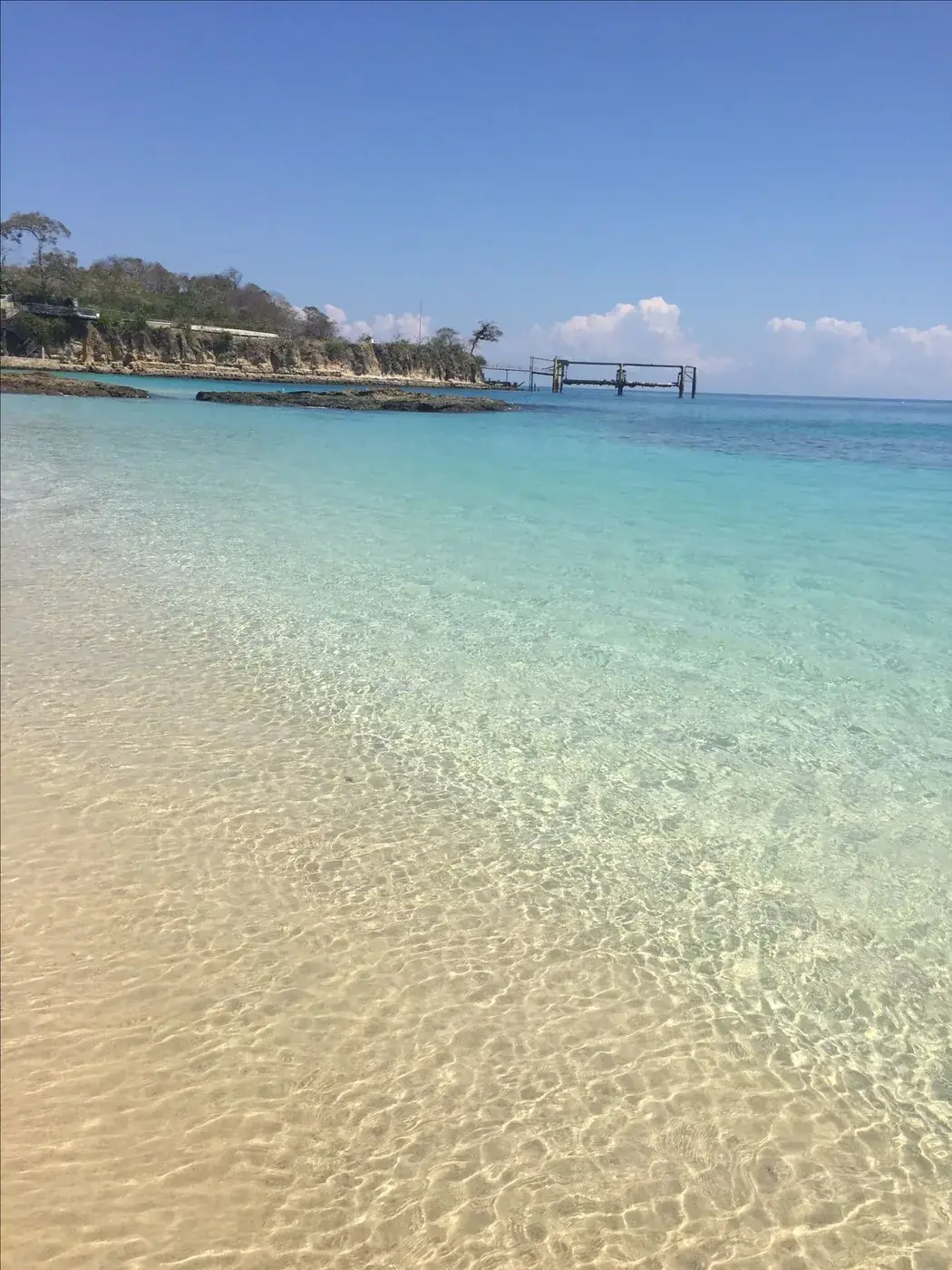 Agua transparente sobre arena clara, con un muelle al fondo