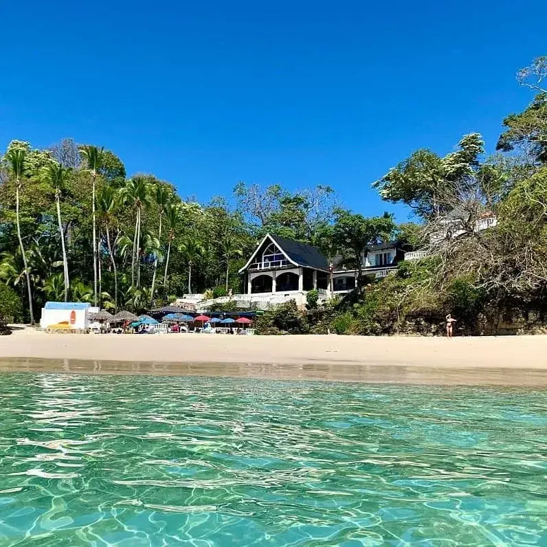 Una playa de arena clara con sombrillas y una casa entre palmeras