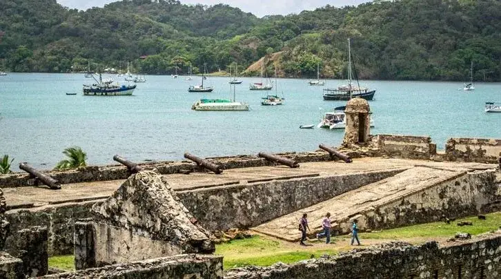 Cañones antiguos sobre la muralla de un fuerte, con veleros en la bahía